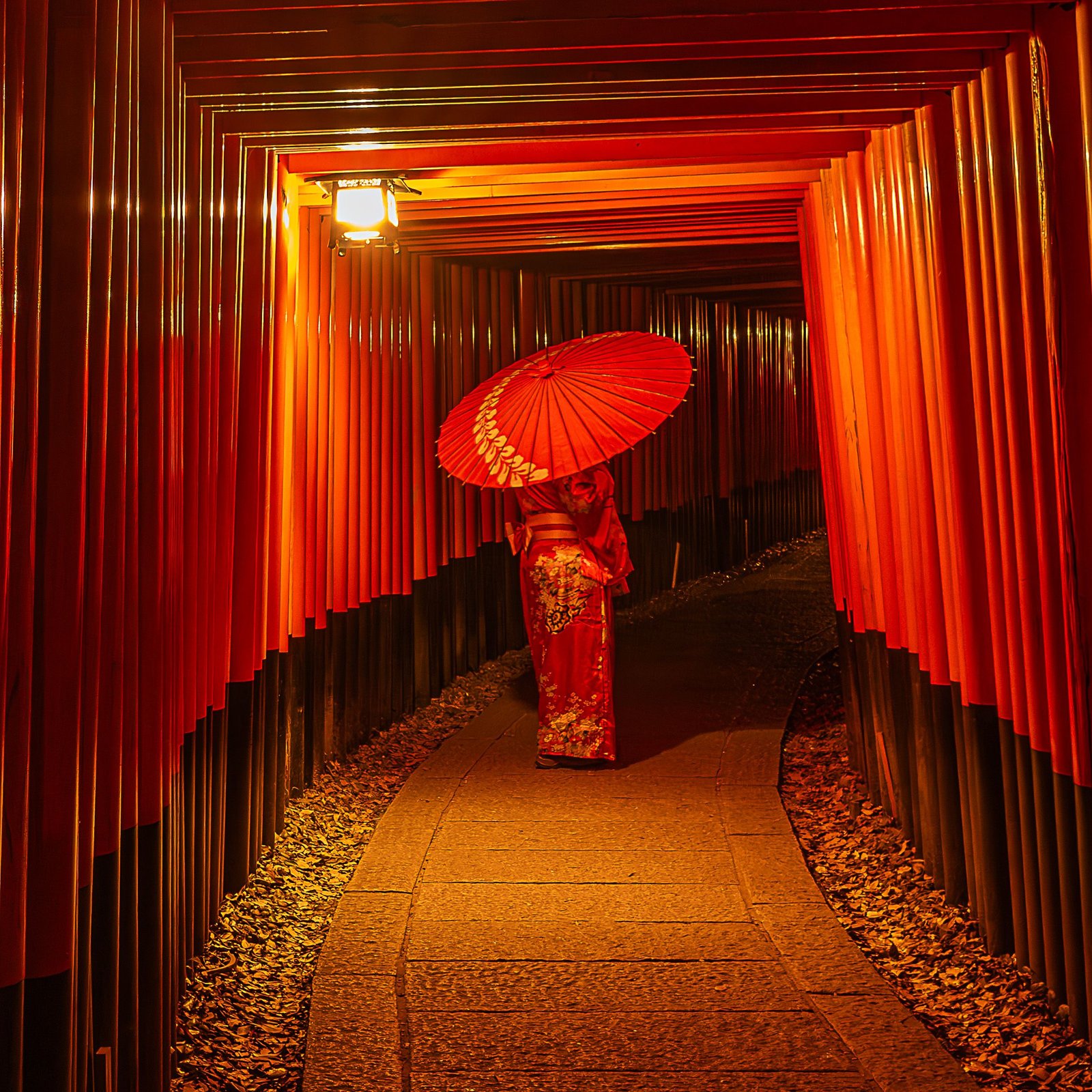 A woman in traditional attire walking with an umbrella under vibrant torii gates at night.