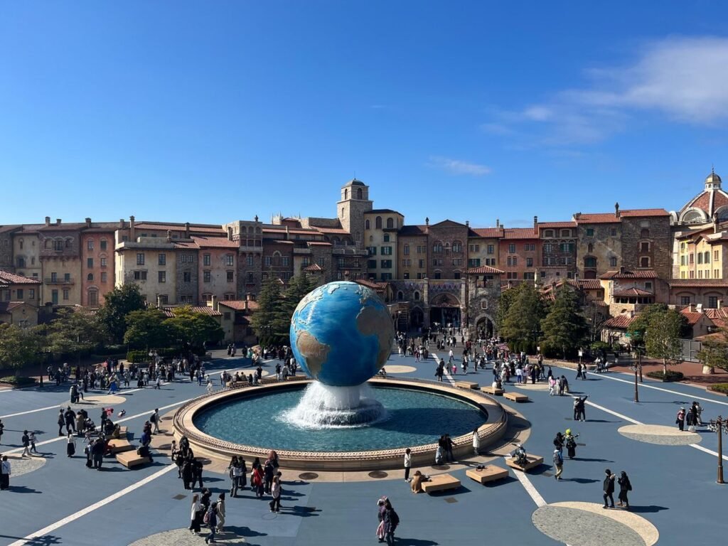 Wide view of a colorful piazza with a central globe fountain, bustling with people under a clear blue sky.