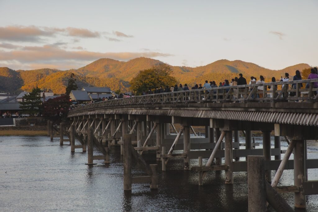 Crowd of unrecognizable travelers walking on aged wooden Togetsukyo Bridge crossing river and leading to Kyoto district near mountains against cloudy sunset sky