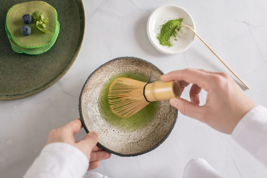 Top view of anonymous cook mixing green food coloring with water using whisk on white table with pancakes